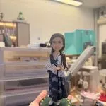 手掌托起的場景公仔：角色穿白色上衣搭配深藍花卉紋長裙，長髮披肩並手持白底花紋折扇；腳踩綠色圓形底座，周圍點綴彩色花瓣碎片，前方白色小銘牌寫有「YAN」。背景為室內工作台與收納抽屜。
