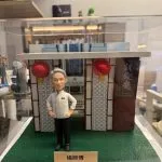透明展示盒中的場景公仔：身穿白色主廚外套與黑色長褲的角色雙手叉腰站在人造草地質感底座上，背後為帶幾何格紋的餐館門面與兩盞紅色燈籠，右側有直立看板印有店名中英文；前方米色銘牌以中文寫有稱呼與「榮休」祝賀語。