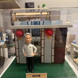 透明展示盒中的場景公仔:身穿白色主廚外套與黑色長褲的角色雙手叉腰站在人造草地質感底座上,背後為帶幾何格紋的餐館門面與兩盞紅色燈籠,右側有直立看板印有店名中英文;前方米色銘牌以中文寫有稱呼與「榮休」祝賀語。