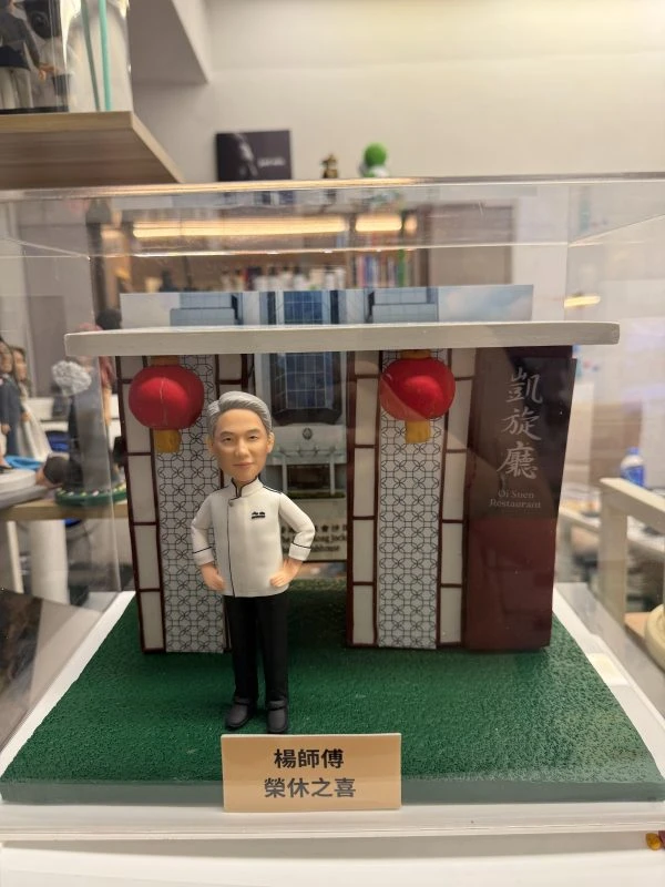 透明展示盒中的場景公仔：身穿白色主廚外套與黑色長褲的角色雙手叉腰站在人造草地質感底座上，背後為帶幾何格紋的餐館門面與兩盞紅色燈籠，右側有直立看板印有店名中英文；前方米色銘牌以中文寫有稱呼與「榮休」祝賀語。