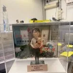 透明展示盒內的場景公仔：戴圓框眼鏡的角色抱著戴紅色蝴蝶結的小貓，腳下為灰色石板紋小底座，前方米色銘牌以英文寫有稱呼、部門縮寫與「Happy Retirement」等字樣（此處不逐字轉錄）；背板印有康樂及文化相關單位與地區名稱的中英文標識與圖案。