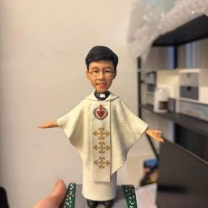 手掌托起的場景公仔:角色穿白色披肩式禮服,胸前有紅色聖心圖樣與垂直金色刺繡花紋;雙臂微展、腳穿黑鞋;站在灰色石板紋底座上,兩側綠色邊條點綴小花。前方銅色銘牌寫有「Ordination blessings for Fr. Benedict Shum 13 • 6 • 2026」。背景為室內書架與收納櫃。