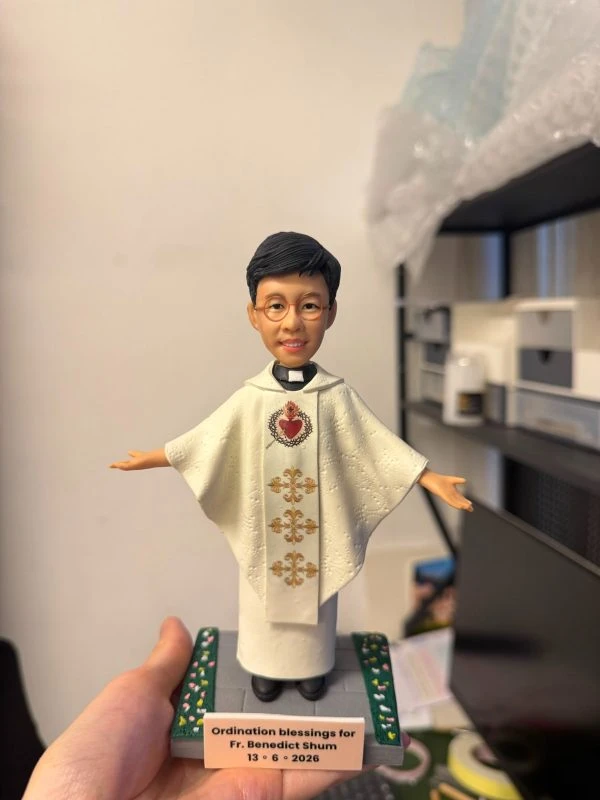 手掌托起的場景公仔：角色穿白色披肩式禮服，胸前有紅色聖心圖樣與垂直金色刺繡花紋；雙臂微展、腳穿黑鞋；站在灰色石板紋底座上，兩側綠色邊條點綴小花。前方銅色銘牌寫有「Ordination blessings for Fr. Benedict Shum 13 • 6 • 2026」。背景為室內書架與收納櫃。