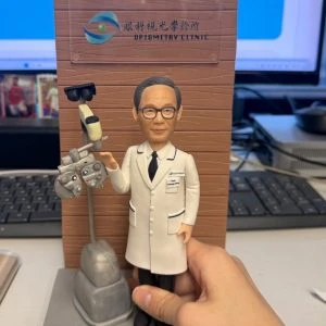 場景公仔立於灰色底座:角色戴黑框眼鏡、穿白色醫師袍與黑色領帶,旁邊有驗光儀器(phoropter)與檢查燈裝置;背後為木紋直立背板,印有眼科/驗光診所的中英文標識;前方桃色銘牌以英文寫有感謝語(此處不逐字轉錄);背景可見鍵盤、螢幕與桌面,右側有手扶住底座。