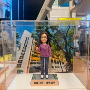 透明展示盒中的場景公仔：角色戴黑框眼鏡、穿紫色針織上衣（胸前有月桂環圖樣）、藍色牛仔褲與白鞋；站在灰色石板紋底座上，兩側綠邊點綴小花。前方淺色銘牌寫有「春風化雨，桃李滿門」。背板為校園大樓與綠樹景色，左側立柱可見多行中文校名文字。