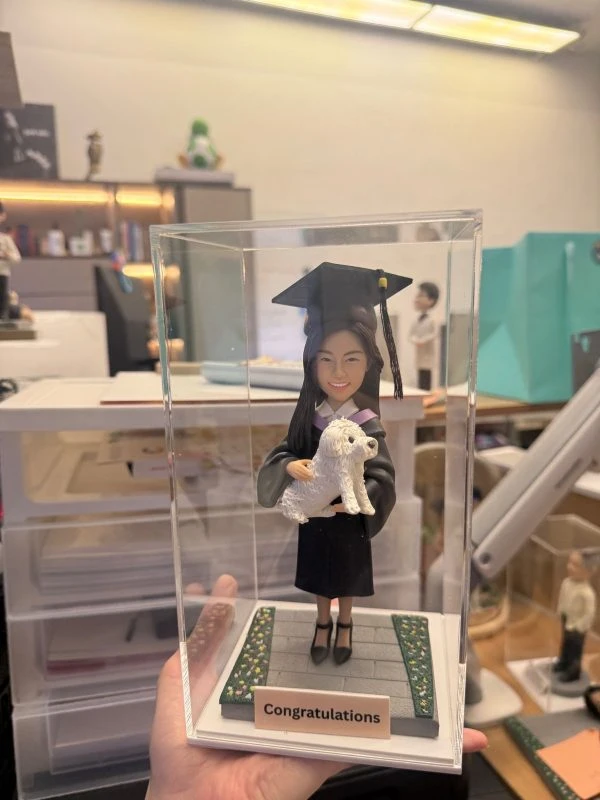 透明展示盒中的場景公仔：角色穿黑色學袍配淡紫領緣、戴學士帽與流蘇，懷抱一隻白色小型犬；站在灰色石板紋底座上，兩側綠邊點綴小花。前方淺色銘牌寫有「Congratulations」。背景為室內工作桌與收納櫃。