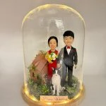 玻璃罩中的婚禮場景公仔：新娘穿紅禮服手捧黃白花束，新郎著黑色西裝配紅色領結與口袋巾；前方有一隻黑白相間的貓站立，其後為小木屋造型與綠植苔面。木質圓形底座周圍繞有暖色燈串，前置金色銘牌寫有「Happy Wedding」。