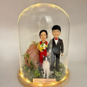 玻璃罩中的婚禮場景公仔:新娘穿紅禮服手捧黃白花束,新郎著黑色西裝配紅色領結與口袋巾;前方有一隻黑白相間的貓站立,其後為小木屋造型與綠植苔面。木質圓形底座周圍繞有暖色燈串,前置金色銘牌寫有「Happy Wedding」。