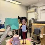 手掌托起的場景公仔：角色戴黑色軟呢博士帽、穿黑底紅邊的學術禮袍，雙手持一張白色證書；旁邊擺放粉色雙層慶祝蛋糕，裝飾有珍珠球與立體花朵，圓牌貼有英文字樣與日期；公仔與蛋糕站在淺黃色磚紋平台上。背景為室內工作室。