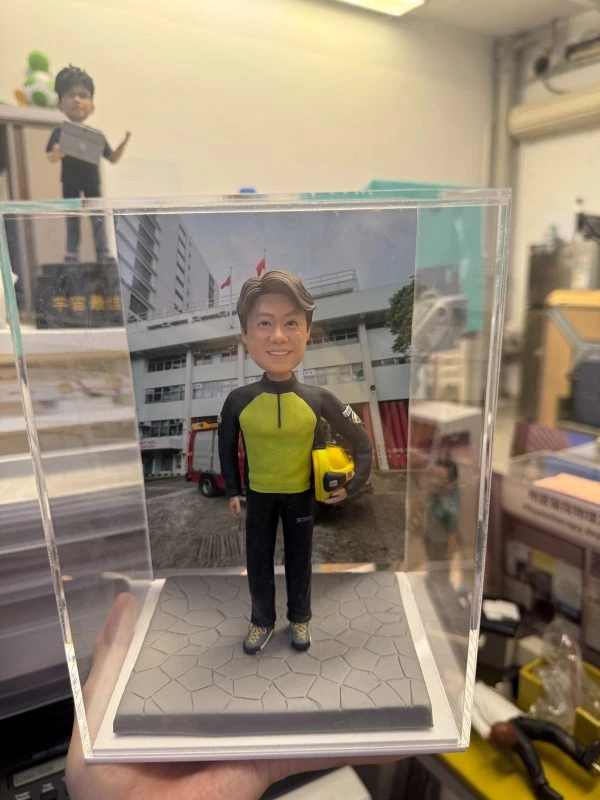 透明展示盒中的場景公仔：角色穿螢光黃綠色上衣搭配黑色長袖與長褲，腳穿運動鞋，右臂夾著黃色安全帽；站在灰色六角石板紋底座上。背板是一棟白色多層建築，屋前廣場停有紅色消防車並掛有旗幟。