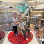透明展示盒中的雙人場景公仔：一位角色坐在淺色藤編椅上，懷抱成疊書本；另一位角色站在身後、手搭肩膀，穿碎花上衣與黑褲；坐者穿灰色上衣與深色長褲、運動鞋。兩人站坐於紅色愛心形底座，邊緣點綴彩色小圓片，前方有紅色愛心標牌寫著「Love」。
