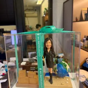 透明展示盒內的場景公仔:角色穿黑色套裝與長靴,手持藍色護照與登機證樣式卡片,另一手拎購物袋與藍色手提包;身後有藍白相間的地球造景與小型沙灘地面。左側是辦公桌(上放筆電、文件、咖啡杯)與一疊紙箱;桌前標示「NVDA $1000」字樣。展示盒外綁有綠松石色緞帶印有「CUTEFIGURE HK」。背景為室內辦公空間。
