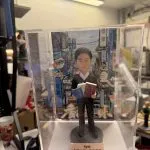 透明展示盒中的公仔：角色穿黑色西裝白襯衫，雙手各抱一本藍、棕色書本；腳下為灰色圓形底座。背板是色彩繽紛的城市街景與大型廣告牌。前方銅色小銘牌黑字寫有「Kyle」與中文生日祝福語。