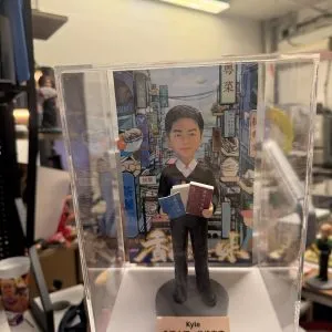 透明展示盒中的公仔:角色穿黑色西裝白襯衫,雙手各抱一本藍、棕色書本;腳下為灰色圓形底座。背板是色彩繽紛的城市街景與大型廣告牌。前方銅色小銘牌黑字寫有「Kyle」與中文生日祝福語。