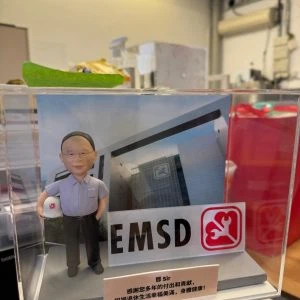 透明展示盒內的場景公仔：角色戴灰色短髮、穿淺灰襯衫與深色長褲、黑鞋，左手握一頂白色安全帽（側面印紅色扳手標誌）。背板為現代感玻璃外牆建築，上貼有同款圓角紅底白色工具標。前方立有大型「EMSD」字樣與紅色工具圖示的立體牌。底部灰色平台前有淺色銘牌，寫著繁體中文感謝賀詞與「鄺 Sir」稱呼。