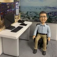 一個穿淺藍色恤衫和卡其色褲的 Q版真人公仔坐喺辦公椅上，前方是擺滿電腦嘅書枱，背景是香港維港風景，適合送俾金融從業員嘅訂造人像公仔禮物。