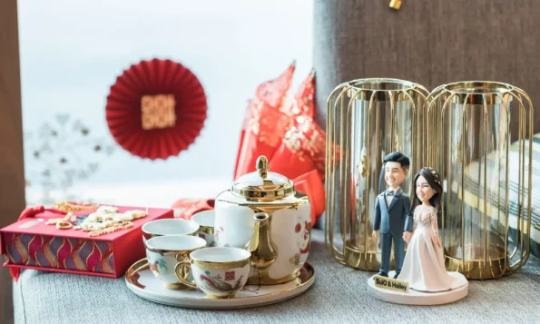 一張充滿藝術感的結婚公仔禮物照片。圖中是為新人SuiO和Hailey度身訂造的結婚公仔，他們穿著西裝和婚紗手牽手，擺放在雅緻的金色家品前。這款精緻的結婚公仔訂造作品，是充滿心思的新人結婚禮物、送好友結婚禮物或結婚一週年禮物的理想選擇。