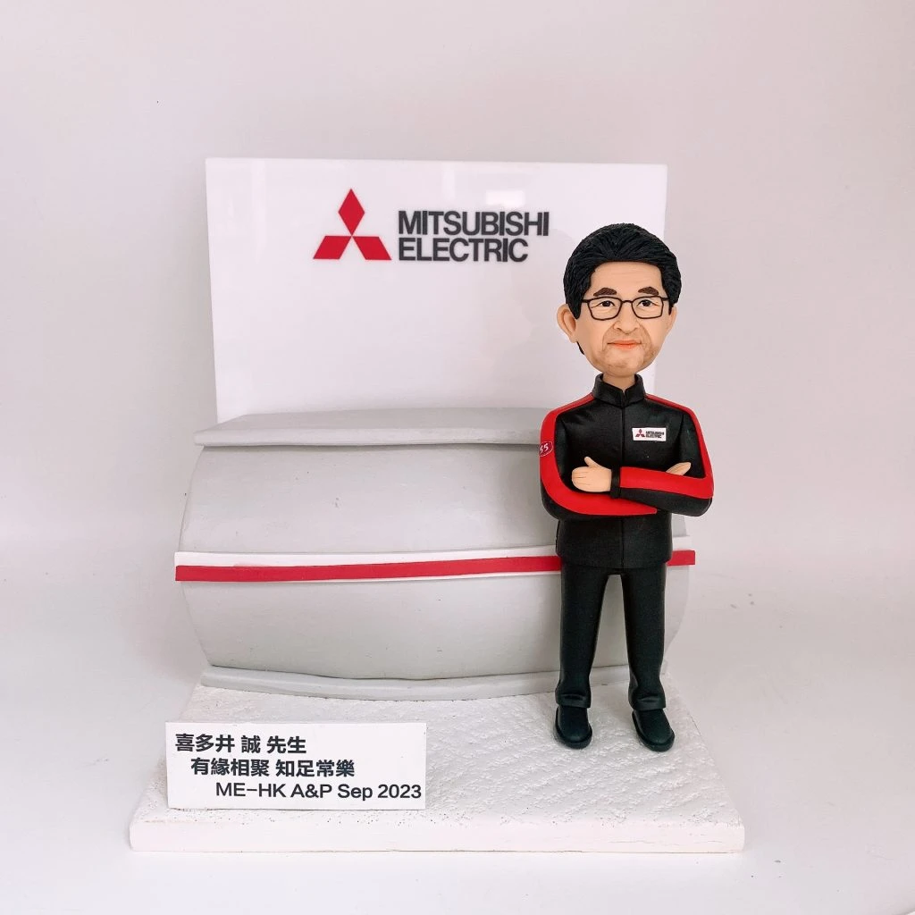 穿著Mitsubishi Electric制服的男性人像公仔,雙手交叉站立,背景有公司標誌和展示牌。