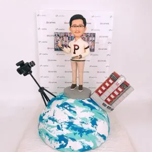 新聞發佈會主持人主題人像公仔：短髮戴眼鏡的男士站在藍白相間的地球造型底座上，身穿米色上衣與卡其長褲，左手比出講解手勢、右手持平板；身旁有黑色攝影機與三腳架、傾斜的紅灰色高樓模型作為城市意象，背後是印有品牌字樣與人物合照的發布會背板。整體呈現新聞舞台與城市連結的概念場景，適合作為新聞發佈會主持人人像公仔榮休禮物與紀念展示。
