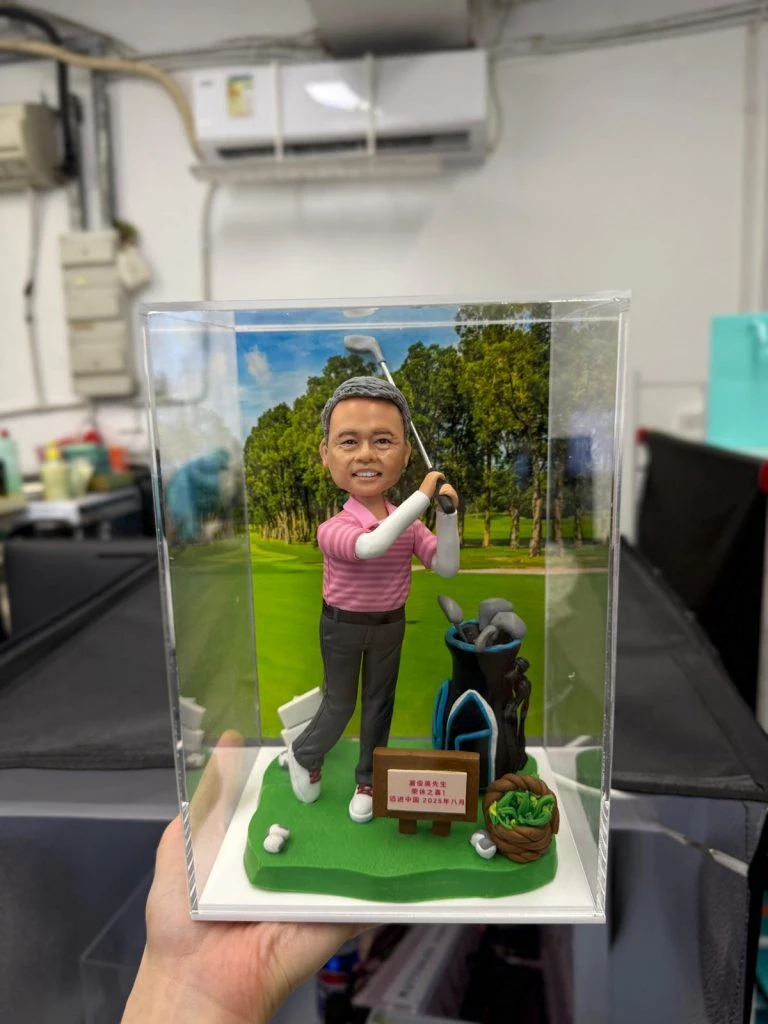 【老闆退休】哥爾夫球主題人像公仔：中年男士公仔穿粉紅條紋高爾夫上衣、深灰長褲與白球鞋，雙手完成揮桿動作站在綠色草地底座上；身後有黑藍相間的球桿袋與多支球桿，腳邊散落白色高爾夫球，前方木牌寫有祝賀與日期字樣。背景為樹林環繞的球道照片板；整體置於透明防塵盒內，呈現「老闆退休」慶賀與休閒品味的哥爾夫球人像公仔。
