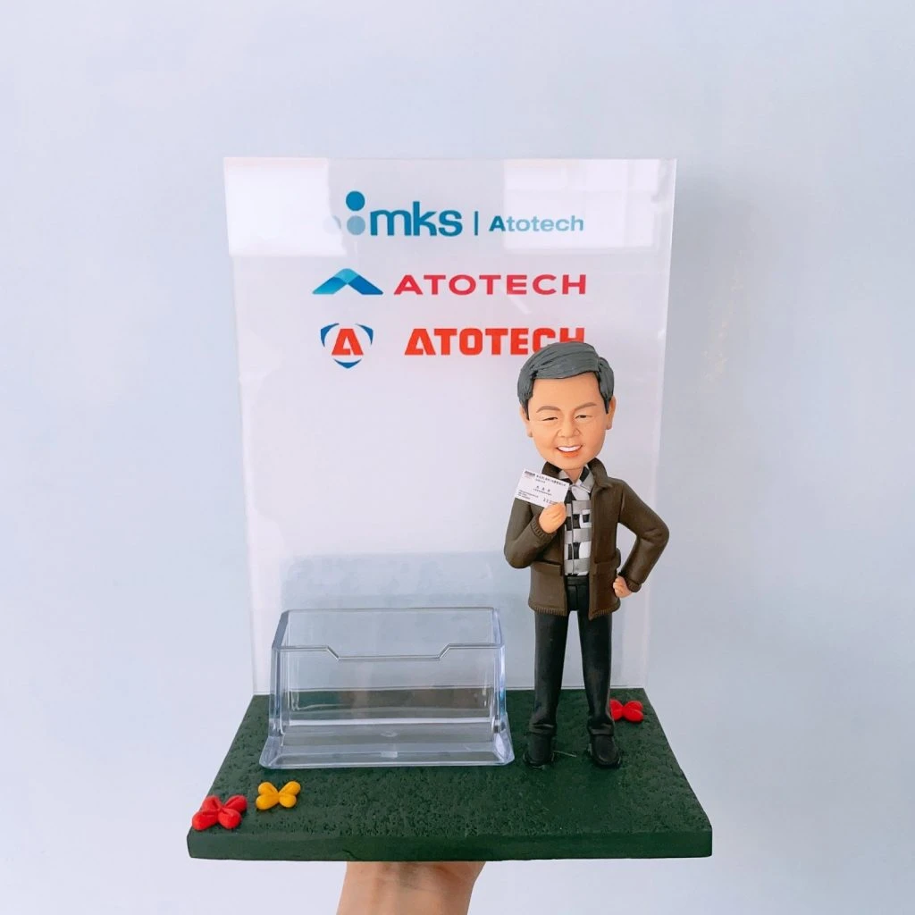 一座結合名片卡片座的商務風人像公仔擺設:灰髮微笑男子穿深色外套、格紋襯衫與黑色長褲,右手持名片、左手叉腰,站在深綠色長方形底座上;底座前緣點綴紅、黃小花。中前方配置透明塑料卡片座,可放置名片或卡片。背板為透明壓克力板,上方印有「mks | Atotech」與多個「ATOTECH」字樣與圖形標記。整體呈現乾淨俐落的辦公室桌面收納與展示效果,具紀念與實用功能。