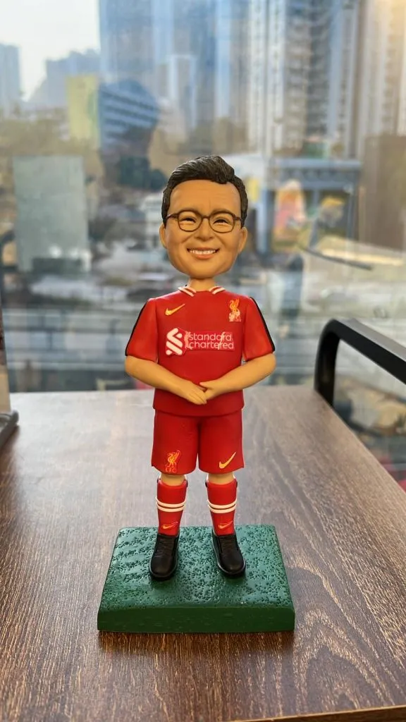 一個穿著紅色利物浦足球隊球衣的人像公仔，面帶微笑，站在綠色草地底座上。公仔穿著利物浦球衣，帶有俱樂部的標誌、Nike標誌，並佩戴紅色的球襪和黑色球鞋。背景為模糊的城市景觀，突出顯示球員的專屬紀念樣式。
