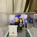 一款客製化牧師人像公仔，呈現一位牧師與女同工站在學校講台後，背景是校園建築，講台有十字架與校徽，是紀念牧師在校服事的榮休感謝禮物。