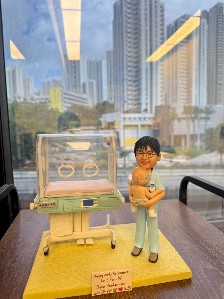 訂製兒科醫生人像公仔｜醫護人員穿著淺藍色醫療制服，懷抱嬰兒，旁邊放置新生兒保溫箱（嬰兒培養箱），細節包括透明罩與觀察孔，展現新生兒科醫護的專業環境，底座刻有退休祝福字句，作為醫生退休禮物極具紀念價值。