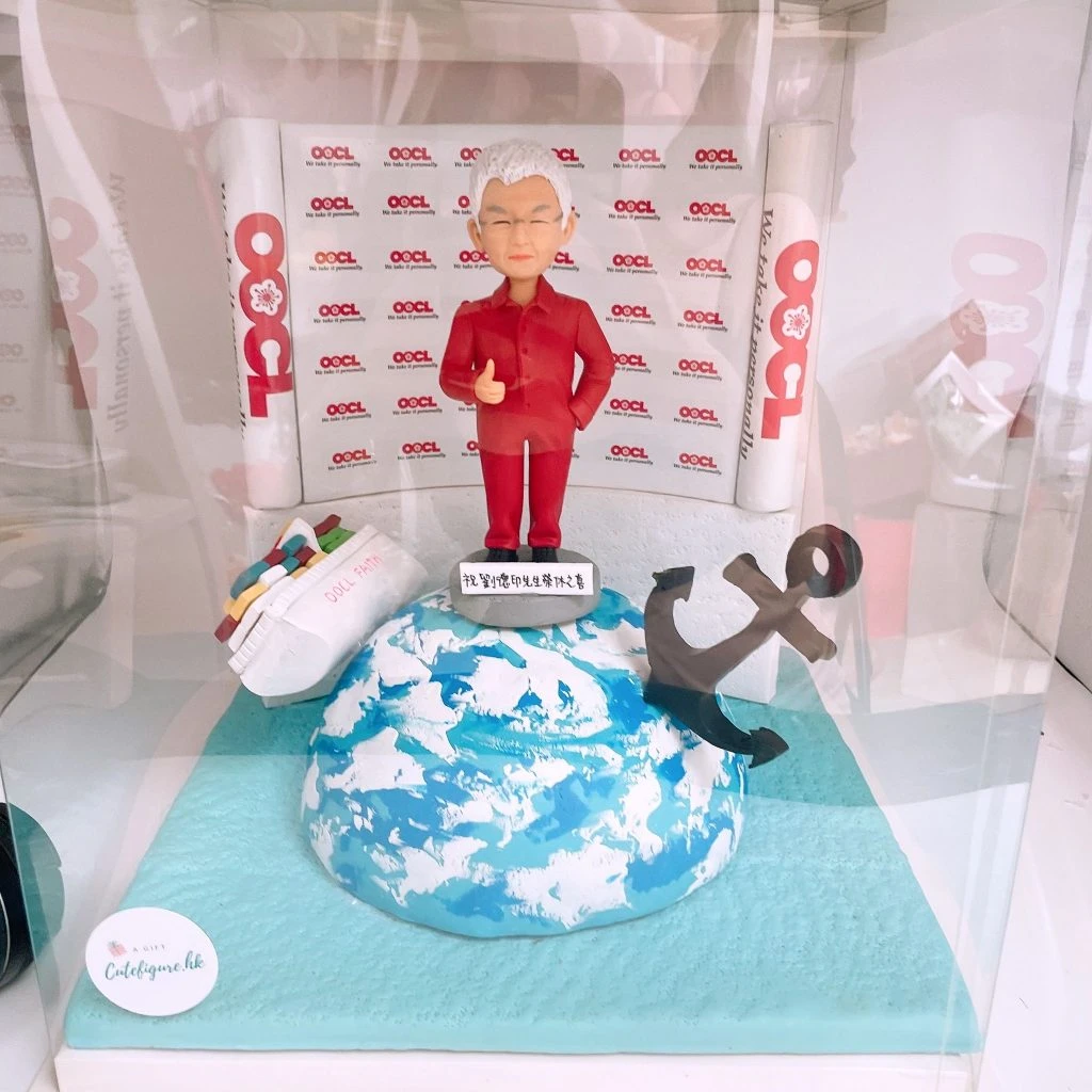 航運主題老闆退休禮物人像公仔：銀白短髮男子穿紅色中山裝風格套裝，微笑比讚站在小圓台上；腳下為藍白相間的地球海面造型底座。左側是一艘載滿彩色貨櫃的白色船模型（側寫有英文字），右側立有黑色金屬感錨模型；背板與兩側圓柱裝飾反覆印有「OOCL」字樣與圖案。整件作品置於透明展示盒中，象徵航運生涯圓滿謝幕的紀念擺設。