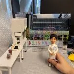 訂製醫生人像公仔擺設｜醫護人員穿著白袍站立微笑，背景為「香港兒童醫院」(Hong Kong Children’s Hospital)，旁邊配有實驗室場景，包括顯微鏡、培養瓶及實驗桌，細緻還原醫學研究環境，是專屬醫生的退休禮物或紀念禮品。