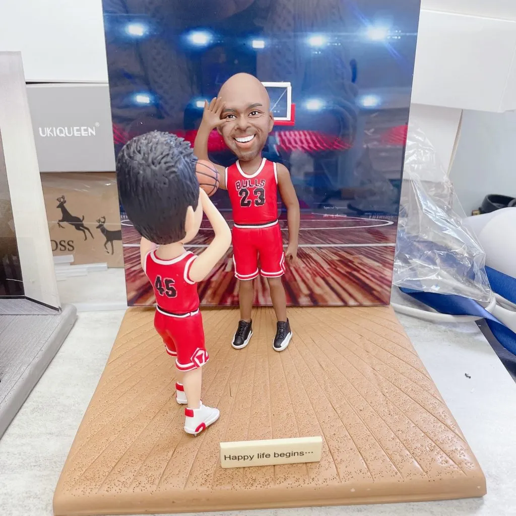 籃球場景雙人籃球人像公仔：一位身穿紅色23號球衣的籃球明星造型公仔站在籃框前微笑致意，另一位穿紅色45號球衣的年輕球迷公仔在前景持球仰頭準備投籃；木紋球場地台附有金色小銘牌寫著「Happy life begins…」，背景是燈光明亮的室內球館與籃板框，整體呈現偶像與粉絲在球場互動的溫馨畫面。