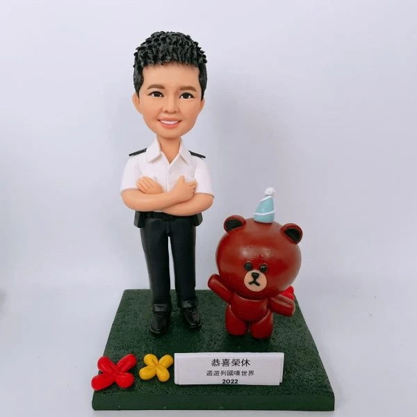 一款為香港紀律部隊人員訂製的榮休人像公仔，主角身穿白色制服，身旁站著一隻戴派對帽的Brown熊大卡通公仔作為背景配件裝飾。