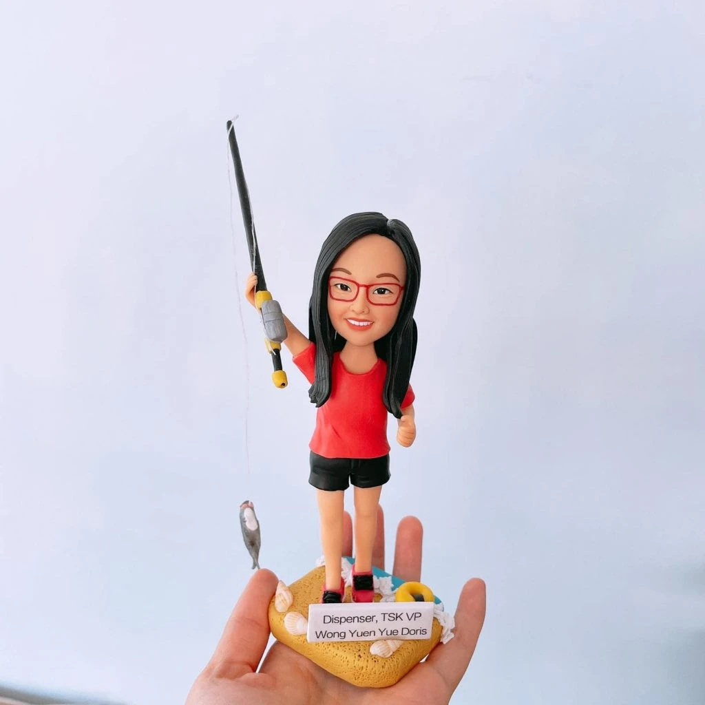 一款女性釣魚主題的人像公仔站在海灘風底座上：長直黑髮、紅色眼鏡，穿紅色上衣與黑色短褲、紅黑運動鞋。右臂高舉黑灰相間的釣竿，細釣線垂下，左側懸著小魚模型；底座有貝殼、救生圈等沙灘元素，前方貼有名牌「Dispenser, TSK VP Wong Yuen Yue Doris」。