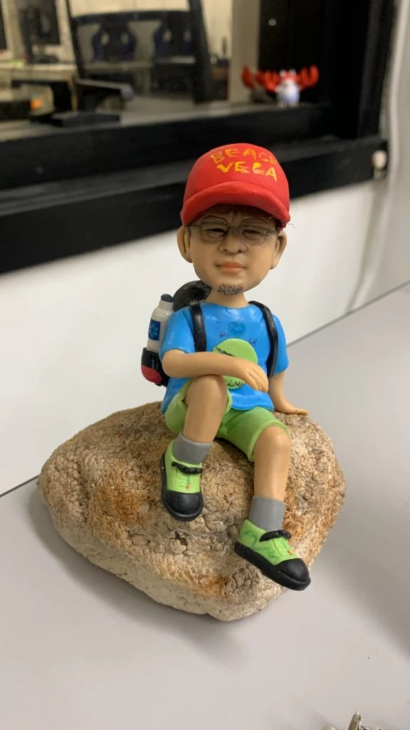 行山主題人像公仔：戴紅色帽子與眼鏡的男子公仔身穿藍色機能上衣、淺綠色短褲與灰色中筒襪，腳踩螢光綠黑色登山鞋，背負藍黑登山背包（側邊掛水瓶），坐在一塊帶有粗糙紋理的淺褐色岩石上休息；整體呈現輕裝郊遊與登山途中小憩的氛圍，適合熱愛行山者的客製人像公仔展示擺設。