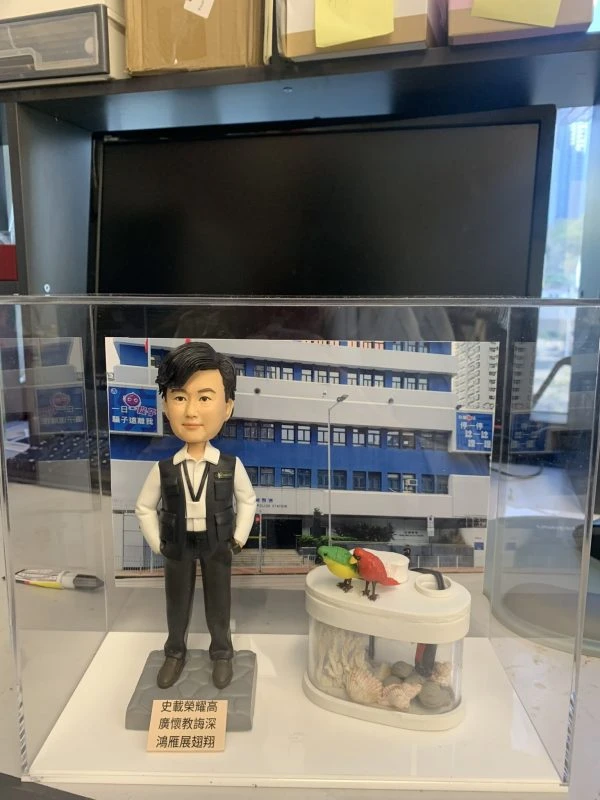 一款為香港警務人員訂製的晉升人像公仔，主角為一名身穿戰術背心的警官，旁邊配有一個帶愛情鳥的魚缸配件裝飾，背景為警署照片。