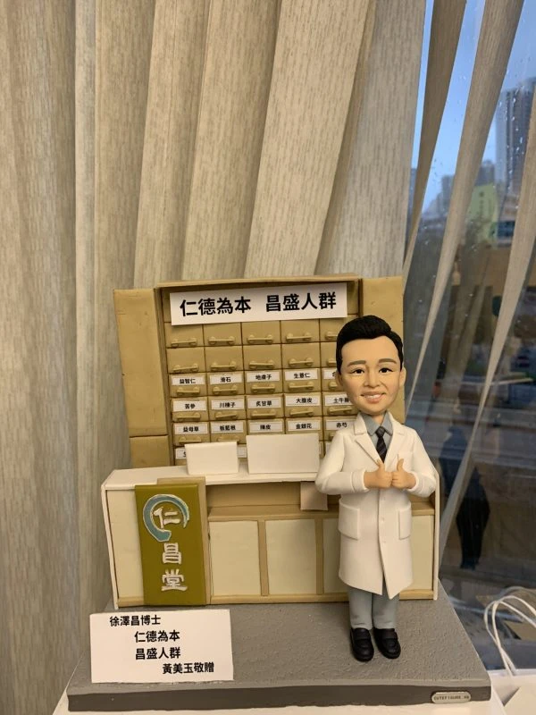 中醫師人像公仔，身穿白袍站立於中藥櫃前，背景是傳統中藥藥材抽屜櫃，上方寫有「仁德為本 昌盛人群」的牌匾。此客製化中醫公仔細節逼真，展現醫者仁心與專業精神，是送給中醫師的獨特禮物與紀念品。