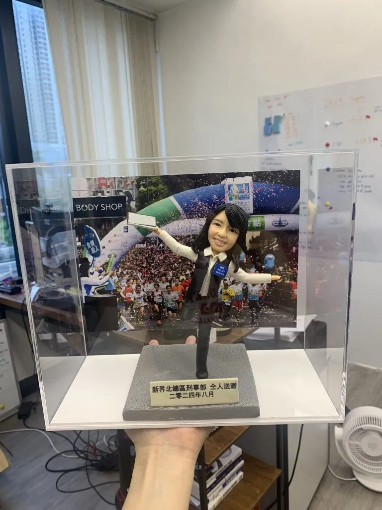 以手作定格高光時刻:這款警察人像公仔採用動態展臂站姿與單腳微踮設計,臉部眉眼弧度、嘴角微笑、髮絲刻線和背心/襯衫的摺痕都逐一刻畫。背板選用大型活動現場影像,與透明展示盒共同營造儀式感;灰色質感底座與金色牌匾則強化典藏層次。無論作為升遷誌慶、團隊紀念,或致謝同事的專屬送禮,皆能體現專屬故事與心意。