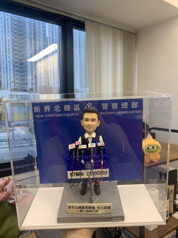 精緻手工製作的警務人員人像公仔（人像公仔、警察人像公仔），置於透明亞加力展示盒內；公仔五官雕刻細膩，穿白色恤衫、黑色戰術背心、黑褲與黑鞋，站在灰色石紋底座上。前方設多支新聞咪高峰連不同媒體咪牌，咪線以紅、藍、黑線纜呈現；中央有編號牌「NTNRN 22000502」。底座金屬牌刻字「新界北總區刑事部 友人送贈 二零二四年八月」。背板印有「新界北總區 警察總部 NEW TERRITORIES NORTH REGIONAL HEADQUARTERS」字樣；背景為室內辦公環境與窗外高樓，右側可見毛絨公仔擺設。整體做工精美、塗裝乾淨，還原警方記者會發言場景。