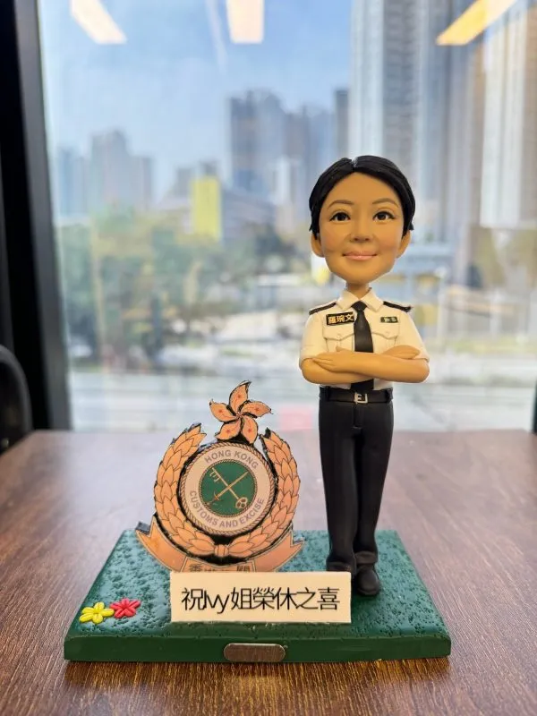 「香港海關人像公仔，身穿白色制服襯衫與黑色長褲，雙手交叉胸前，神情自信專業。底座展示『Hong Kong Customs and Excise』徽章，以及『祝 Ivy 姐榮休之喜』字樣，象徵此公仔為慶祝香港海關職員榮休（退休）特別訂製的專屬紀念禮物。此公仔細節精緻，適合作為榮休禮物、警察或海關紀念品，以及專屬人像公仔收藏。」