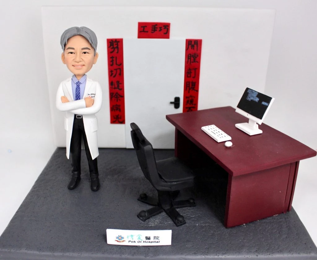訂製醫生人像公仔，角色穿著白色醫生袍，雙手交叉站在辦公桌旁，桌上有電腦螢幕、鍵盤與滑鼠，背景寫有醫療對聯「剪扎切縫除病災、開膛訂腹癒百病」。底座標示「博愛醫院 Pok Oi Hospital」，象徵醫生的專業與醫療貢獻，適合作為榮休、紀念或醫護禮物。