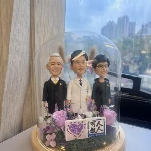 三人造型人像公仔擺設，其中一位身穿白袍象徵醫生，另兩位穿著西裝，公仔被安置在透明玻璃罩內，底座以木質打造並配有乾燥花佈置及燈飾。裝飾上附有「可人教人」「All Are Educable」及「馬錦明慈善基金馬可賢伉儷書院」的牌子，展現獨特紀念與致敬氛圍。此醫生與醫護相關公仔設計可作為醫護禮物、紀念品或專屬收藏。