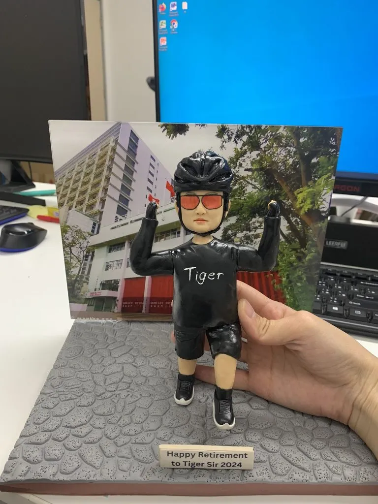 一款為Tiger Sir訂製的退休人像公仔，公仔身穿印有「Tiger」字樣的單車服，做出衝線勝利手勢，背景是消防局照片，底座銘牌寫有「Happy Retirement to Tiger Sir 2024」。