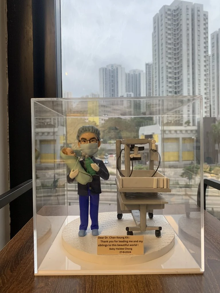 醫生人像公仔模型，身穿手術服與口罩，雙手抱著嬰兒，身旁放置新生兒保溫床與醫療設備，背景為都市高樓景色。此公仔為客製化醫生禮物，適合作為婦產科醫生、新生兒科醫護的感謝紀念品。