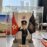 透明展示盒內的警察人像公仔：身著深藍制服與制帽，右手舉至眉間敬禮，左手自然下垂；胸前有肩帶與口袋釦飾，黑色皮鞋。人物站在灰色方形地台中央，左右各有一面旗幟：左側為紅底白花旗，右側為深藍色警徽旗。前方白底名牌印有中英對照的職級稱呼與姓名；名牌左側帶有警徽圖案。背板中央見到圓形紫色標誌，展示盒置於工作桌上，周圍有手工具與模型配件；窗外為高樓景觀。整體呈現正式、榮譽與頒贈性質，適合作為升遷、榮休或部門致意的送禮。