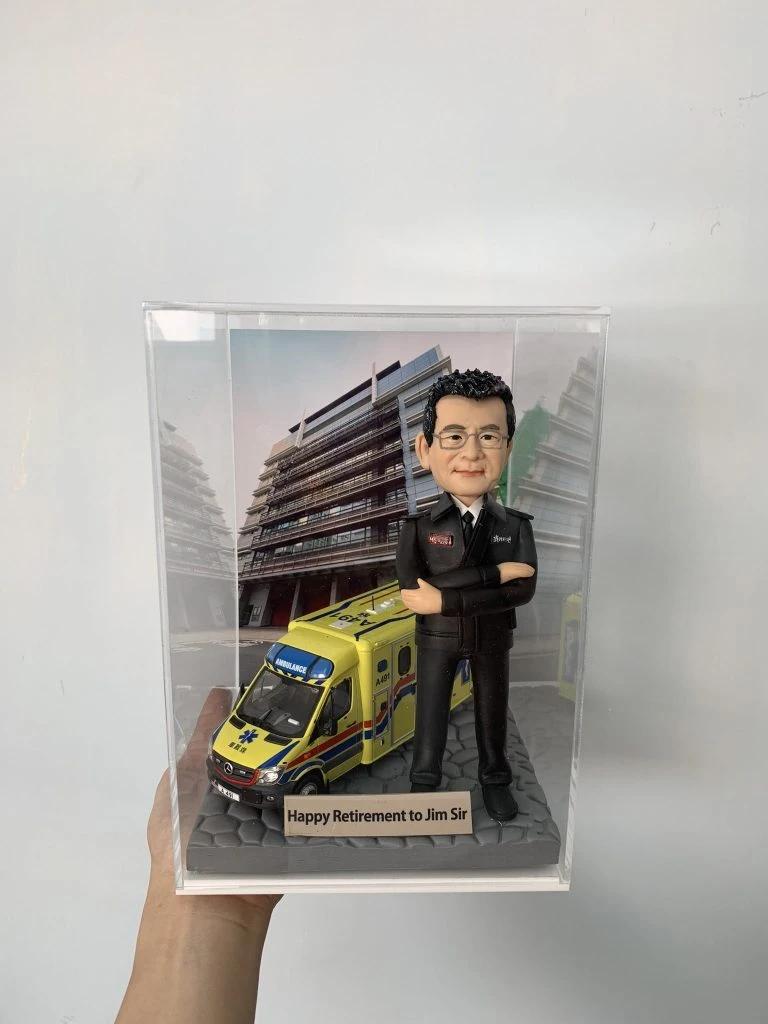 一款為救護員長官 Jim Sir 訂製的退休消防人像公仔，公仔身穿制服，與一輛香港救護車模型一同站立在消防局照片背景前，底座刻有退休祝福語。