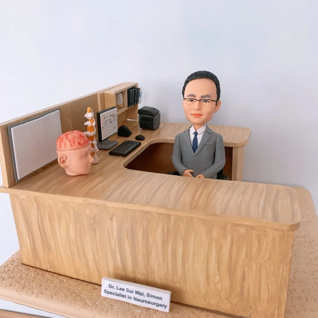 神經外科醫生人像公仔,身穿西裝的男醫生坐在診間辦公桌後方,桌面擺放大腦模型與脊椎模型,象徵專業的神經外科醫學。辦公桌上有電腦、鍵盤、醫學書籍與醫療文件,細緻還原醫生診間場景。此客製化醫護公仔適合作為醫生禮物、醫護紀念品、退休紀念或升職賀禮。