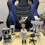 訂製醫生人像公仔：一位穿著白袍的醫生，雙手交叉站立於手術室中央，背景配備手術檯、無影燈、達文西手術機械臂（Da Vinci Surgical System）、監測設備與手術椅，完整還原微創手術場景，展現醫護專業與臨床環境。