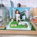 透明展示盒中的醫護人像公仔：一位身穿白袍的女性醫護專業人士，交叉手臂自信站立，右腳踩在一架印有「Cathay Pacific」字樣的小飛機模型上，前方有退休祝賀銘牌；左側可見灰白色的實驗室分析儀模型；背景是威爾斯親王醫院（Prince of Wales Hospital, PWH）的大樓外觀。整體為化學病理科主題的客製禮物。