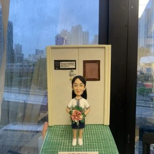 手工訂製醫護人像公仔，人物身穿醫院制服，手持花束，站立於模擬醫院場景模型前。底座刻有「To Miss Sherla Yu, From QMH Neurosurgery team」字樣，顯示來自瑪麗醫院神經外科團隊的致意。此人像公仔是專屬醫護禮物，象徵醫生與醫護人員的專業精神與感謝之情。