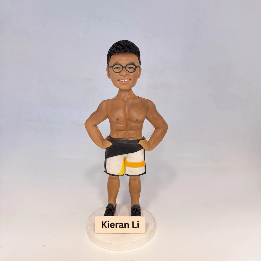 健身主題人像公仔：短髮男子公仔身穿粉紅條紋上衣與深灰長褲、白色運動鞋，雙手做出揮臂動作站在綠色草地造型底座上；旁邊放置黑藍色運動裝備袋與多件器材道具，前方木牌可見客製祝福字樣。背景為綠地與樹林場景的照片板，整體收納於透明展示盒內，呈現健身教練氣質的人像公仔擺設。